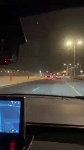 The video shows a night drive on a highway. The view is from the driver's seat, looking forward through the windshield. There are several cars ahead on the road, illuminated by headlights and taillights. Streetlights line the sides of the highway. There appears to be construction or some form of road work occurring, as there are orange cones visible on the left side of the road. The dashboard of the car is visible in the lower portion of the frame, including a digital display screen. Upbeat music is playing in the background.