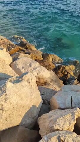 The video shows a close-up, low-angle shot of large, light-colored rocks in the foreground. Behind the rocks, the turquoise ocean water is visible, with small waves crashing against the shore. The water appears clear and shallow near the rocks, with darker water further out. Sunlight illuminates the scene, casting shadows on the rocks. No people or other objects are present.