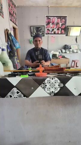 The video shows a man in a barber shop looking into a mirror and holding a smartphone. He is wearing a dark blue short-sleeved shirt and a dark blue kufi. The barber shop appears to be small and somewhat cluttered, with posters of hairstyles on the wall and various barber tools and supplies on a counter. In the foreground, a patterned surface with diamond shapes is visible, likely part of the counter. The man is looking at his reflection and appears to be posing for a selfie or taking a video. He briefly touches his face and then gestures with his hand.