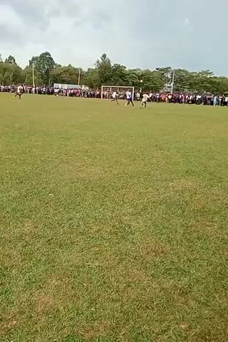 The video depicts a soccer game in progress on a green field, with a crowd of spectators gathered along the sidelines. Several players are visible on the field, engaged in the game. The setting appears to be a local sports ground, with trees surrounding the field and a cloudy sky overhead.