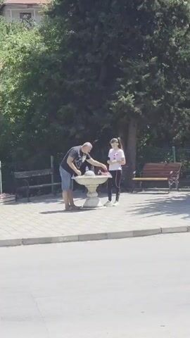 A man and a young woman stand near a decorative fountain in a park. The man, wearing a dark t-shirt and denim shorts, leans over the fountain, appearing to interact with it or something placed on it. The woman, in a white t-shirt, black leggings, and white sneakers, stands to the right of the fountain, holding a pink object. Behind them, park benches are visible, and a large tree provides shade. The ground around the fountain is paved with light-colored tiles, and a paved path or road is in the foreground. The scene is set outdoors on a sunny day.
