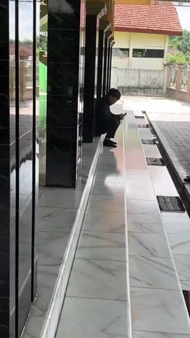 A man in dark clothing sits on a marble step, looking down at his phone. He is positioned in the shaded area of a building's exterior, possibly a mosque, with black tiled columns to his left. The steps are clean and well-maintained, leading down to a walkway paved with patterned stones. Beyond the walkway, a fence separates the area from a building with a red tile roof. A motorbike is parked to the right. The environment appears to be outdoors on a bright day.