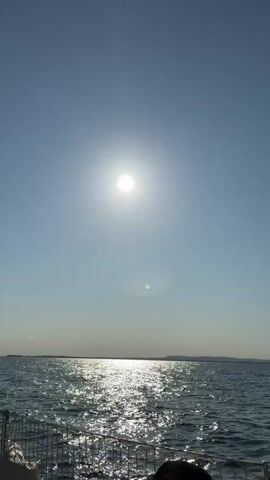 The video shows a bright sun high in a clear blue sky over a calm sea. The sun's reflection creates a shimmering path on the water's surface. In the foreground, the edge of a boat is visible, with some netting and a part of a person's leg. The horizon shows a distant landmass. The camera is pointed upwards, capturing the expansive sky and the sun.