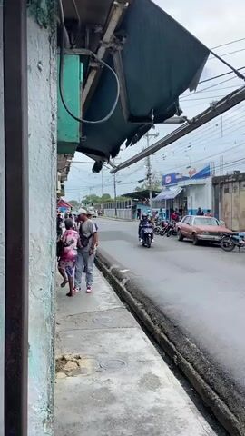 The video opens on a street scene with a tattered awning hanging precariously over a sidewalk on the left. A few people are walking on the sidewalk. A man in a red shirt and a woman in a patterned dress are walking away from the camera. A man in a grey shirt and jeans is walking towards the camera. The street is paved and appears wet. On the right side of the street, there are several motorcycles parked and a few buildings. One building has a Burger King sign. Further down the street, there is a light-colored sedan parked. A person is riding a motorcycle past the parked car. The video pans to the right, showing more buildings and parked motorcycles. A person is walking on the sidewalk in front of a building with a green metal roll-up door. Another person is walking across the street. The camera then focuses on a yellow building with a green door and windows. A person is sitting on a bicycle in front of the building. A man with a white bag slung over his shoulder is walking away from the building. The video then pans to the right again, showing a building with a stone facade and a red awning. Several people are standing outside the building. A person is holding a microphone and speaking. Another building, seemingly under construction, is visible behind the businesses. A red flag is partially visible in the bottom right corner of the frame. The sky is overcast.