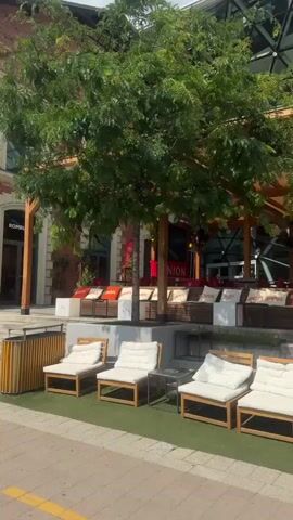 The video shows an outdoor seating area of a restaurant or cafe. In the foreground, there are four white cushioned lounge chairs made of light-colored wood, arranged on a patch of artificial turf. Small black metal tables are placed between the chairs. A paved walkway with a yellow line runs in front of the turf. In the background, a large tree with green foliage provides shade over a covered patio area. This patio features more seating, including cushioned benches and chairs, with various pillows. A sign with the word "BÁZIS" is visible on the building's facade. The building behind the patio has a distinctive modern design with a glass exterior made up of triangular panels. The scene appears to be during daylight hours, possibly a sunny day, given the lighting and shadows. No people are visible in the video.