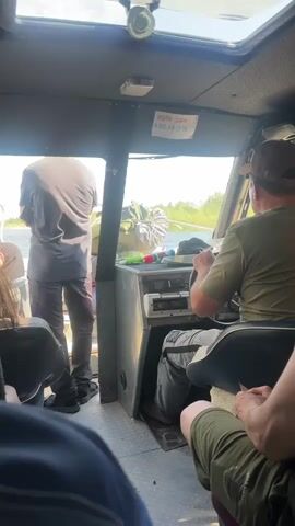 The video shows the interior of a boat, looking forward through a cabin. A person wearing a brown hat and a green shirt is steering the boat. Another person in a grey t-shirt and grey pants is standing near the front of the cabin, facing away from the camera and towards the water. There are other passengers visible in the foreground, also facing forward. The environment outside the boat appears to be a body of water with vegetation on the banks, under a bright sky. The overall impression is of a boat journey in daylight.