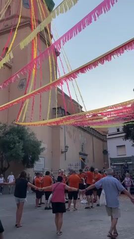A group of people, mostly dressed in orange shirts, are holding hands and dancing in a circle in a town square. They are in front of a large, light brown building that appears to be a church, with a tall clock tower. Colorful streamers in yellow and pink are strung across the sky above the square. The sky is a light blue, suggesting it is either early morning or late afternoon. Other people, dressed in casual clothing, are standing around the periphery of the square, observing the dance. The ground is paved with cobblestones. Some of the dancers appear to be musicians, as one person in the center of the circle is holding what looks like a drum. The overall atmosphere suggests a festive or cultural event.