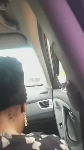 The video shows the interior of a car from the perspective of a passenger. A woman wearing a black headscarf and a patterned shirt is seated in the car, facing away from the camera. She is wearing large hoop earrings. The car's dashboard and side door are visible. The view through the car window shows a blurred outdoor scene with greenery. The video is short and static, with no discernible action or interaction.
