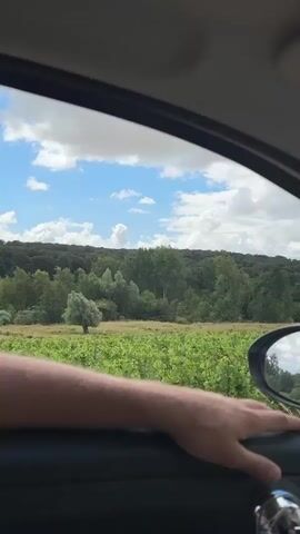 The video captures a view from inside a car driving through a rural landscape. The perspective shifts from looking out the side window to looking through the windshield. We see a person's arm resting on the car door, with their hand visible on the door handle. Outside, a green field, possibly a vineyard, stretches towards a tree-covered hill under a cloudy sky with patches of blue. A large barn filled with round hay bales is visible ahead, situated beside a paved road. The car's interior, including parts of the dashboard and steering wheel, are also visible.