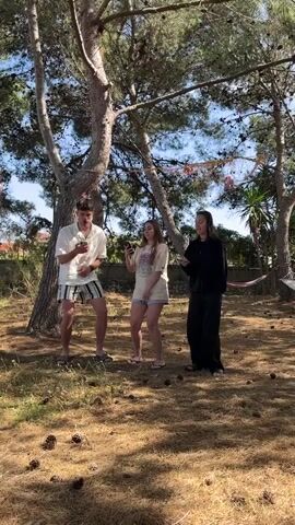 Three young people are standing on dry grass under trees and looking at their phones. The young man on the left wears a white shirt and striped shorts, the young woman in the middle wears a light T-shirt and blue jean shorts, and the young woman on the right wears all black. The ground is covered in dry pine needles and pine cones. In the background, there are trees, shrubs, a fence, and a hammock. The scene appears to be outside in a yard on a sunny day.