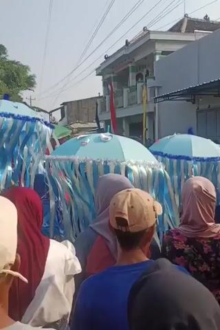 A group of people are gathered outdoors, possibly for a parade or festival. In the foreground, several individuals are seen from behind, wearing headscarves and casual clothing. Dominating the middle ground are large, light blue jellyfish-like props, adorned with white and light blue streamers. These props appear to be carried by people within the crowd. Some of the jellyfish have large cartoonish eyes and a mustache design. In the background, buildings, trees, and power lines are visible. The sky is bright, suggesting a daytime setting. The overall atmosphere appears festive.