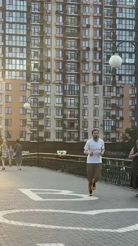 A man with a beard, wearing a white t-shirt and khaki pants, is jogging across a pedestrian bridge. He has on bright orange and yellow running shoes. To his left, a woman in a black and white polka-dot dress is walking and carrying a white bag. Further in the background on the left, another person in a dark outfit is walking away from the camera. The bridge has a decorative metal railing and a paved walkway with white markings.

Behind the bridge, a tall, modern apartment building with many windows dominates the scene. A street lamp with a white spherical light fixture stands near the center of the bridge. In the distance, trees and other buildings can be seen, and a body of water is visible beyond the railing on the right. The sky appears to be a soft orange, suggesting it is either early morning or late evening, with a warm, diffused light. The weather seems pleasant, with no visible rain or harsh conditions.

Visible text includes "МЕТАПТЕКА" and "СТОМАТОЛОГ" on signs attached to the lower levels of the apartment building, indicating a pharmacy and a dental clinic, respectively. The overall atmosphere is that of a calm, urban scene with people engaged in everyday activities.