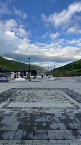 The video opens on a wide-angle, low-angle shot of a paved plaza with a geometric pattern of grey and white stones. In the background, a road curves through a valley flanked by green, mountainous terrain. The sky is a vibrant blue with scattered white clouds, some of which appear dark and heavy.

As the video progresses, the camera pans slightly to the left, revealing a few modern-looking structures. There are white, dome-shaped benches or sculptures scattered across the plaza. A person wearing a blue outfit is visible in the distance, walking away from the camera. Streetlights with a modern design are also present.

Further to the left, more buildings come into view. One appears to be a dark-colored structure with a glass facade, possibly a shop or information center. Another building is dark stone with a visible entrance and windows. More of the white dome-like features are seen. The mountainous landscape continues to dominate the background, with some distant peaks possibly showing snow. The overall impression is of a newly developed public space in a mountainous region. The lighting suggests it is daytime, likely late afternoon given the cloud formations.