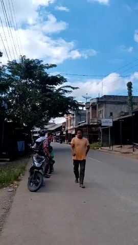 The video shows a street scene in what appears to be a small town in Indonesia. A man in an orange t-shirt walks toward the camera on the right side of the road. On the left side of the road, two men stand near two parked motorcycles.  A third person is seated on the motorcycle closest to the viewer. Another motorcycle drives away from the camera on the right side of the road. Buildings line both sides of the street, with shops and businesses visible. The sky is blue with scattered white clouds. Overhead power lines are visible.