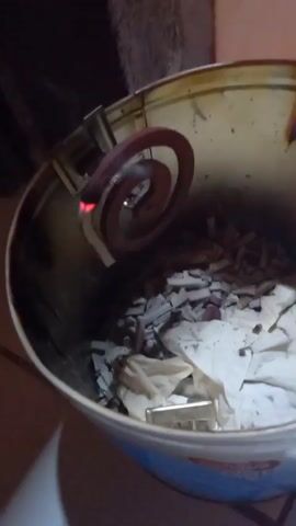 A close-up shot shows a mosquito coil burning inside a metal tin. The coil is suspended by a metal holder attached to the rim of the tin. The tin contains ash and some crumpled paper. Smoke emanates from the burning coil. The scene is dimly lit, suggesting it may be nighttime or an indoor setting with low light.