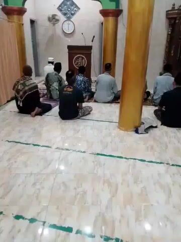 Here is a concise description of the video:

The video shows about ten men sitting on the floor of a mosque, presumably praying. They are all facing a podium with writing on it. The mosque's interior is visible, including pillars, a clock, and tiled flooring. There is a person speaking.