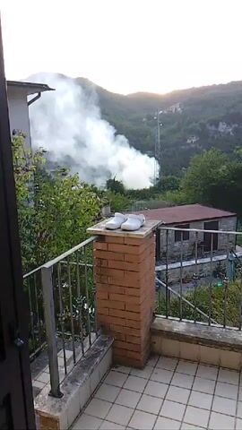 The video opens with a view from a balcony overlooking a hilly, vegetated landscape. A thin plume of white smoke is visible rising from a wooded area on a distant hillside. A tall metal tower with antennas stands on the slope, partially obscured by the smoke. In the foreground, on the balcony, two white shoes rest on top of a brick column. The sky is bright, suggesting it is daytime. The camera pans slightly to the right, revealing a small, rustic building with a red roof and stone walls situated below the hillside. The smoke appears to be a significant size and is drifting upwards and to the left. The environment suggests a rural or semi-rural setting.