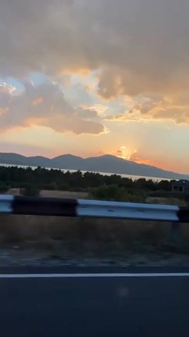 This video captures a scenic view from a moving vehicle during sunset. The camera is positioned to show the landscape passing by. In the foreground, a dark road surface with a white line is visible, along with a black and white guardrail. Beyond the guardrail, there is a stretch of green grass and bushes, transitioning into a line of trees and hills in the mid-ground. The background features a body of water and rolling hills under a dramatic sky filled with clouds illuminated by the setting sun, which is partially obscured by the hills. The warm hues of orange, pink, and yellow paint the sky. A few houses are scattered across the landscape. The movement of the vehicle creates a sense of motion, with the trees and landscape appearing slightly blurred. There are no people or animals visible in the video.
