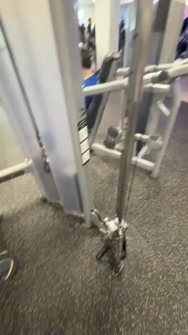 The video shows the interior of a gym. In the foreground, there is a piece of gym equipment with a pulley system. To the left, a person wearing a blue shirt and headphones sits on a weight machine. In the background, there are treadmills, other weight machines, and several people exercising. The gym has purple and pink lighting. The floor is made of black rubber. The walls are light colored. There are mirrors on the walls and a ceiling with acoustic tiles and fluorescent lighting. The video pans to the right, showing more gym equipment and people.