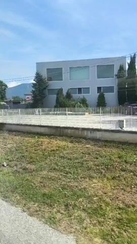 The video appears to be taken from a moving vehicle. In the foreground, there is a grassy area and a paved surface, likely a road or sidewalk. In the middle ground, a white fence separates the grassy area from a blue metal fence. Behind the blue fence, a sign reads "TIR-CAR" in large letters, with smaller text below. A white building with large windows is visible in the background, as well as trees and vegetation. The sky is blue with some clouds, and mountains are visible in the distance. The overall impression is of an outdoor environment, possibly a roadside commercial area.
