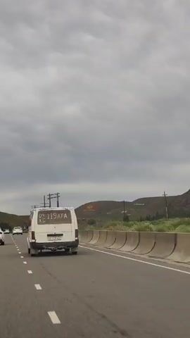 A white van with license plate 09 119 AFA KC is driving down a road. The van has a number written on the back window. There are other cars on the road and power lines on the side. The sky is cloudy. The landscape is hilly.