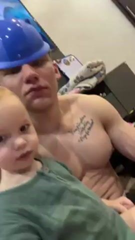 A shirtless man with a blue plastic helmet sits holding a toddler in a green shirt. The man has a tattoo on his upper chest. The toddler looks at the camera and the man looks off into the distance, then down at the toddler. The toddler reaches for the camera and the video ends. In the background there is a TV and various household items. The lighting is bright.