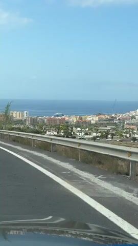 The video captures a roadside view from a moving vehicle, looking out over a coastal city. A bright blue sky with scattered white clouds dominates the upper portion of the frame. Below the sky, a clear blue ocean stretches across the horizon. The city, with its colorful buildings and palm trees, is situated between the ocean and a hilly landscape. A highway guardrail runs along the side of the road in the foreground, and the asphalt road with white lane markings is visible. The vehicle appears to be traveling uphill, providing a panoramic view of the urban sprawl and the sea. There are no people visible in the video. The overall impression is one of a scenic drive along a coast on a sunny day.