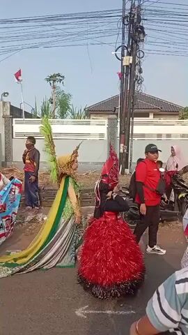 A person in a green and gold elaborate costume with large feathered wings and a long train stands on a street. To their right, another person is dressed in a red, feathery costume with a matching headpiece. A man in a red shirt and black pants, wearing a black cap, stands behind the person in the red costume. A woman in a headscarf is visible further back. Several motorcycles are parked on the side of the road. A small child in a striped shirt is in the foreground, facing away from the camera. A building with a white fence is in the background. Wires and utility poles are prominent in the upper portion of the frame. A small Indonesian flag is flying from a pole to the right of the building. The scene appears to be a street performance or parade.