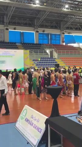 The video shows a dance competition in a gymnasium. Many young women are lined up, dressed in glittering dance costumes. They appear to be participants, and some are holding signs with team names. A few men are also present, some in more formal attire, possibly coaches or organizers. In the foreground, a few people are seated at tables, possibly operating recording equipment or managing the event. The environment includes bleachers for spectators in the background, a large banner with event information, and a polished wooden floor typical of a sports venue. The overall atmosphere suggests an organized event with a focus on dance performance.