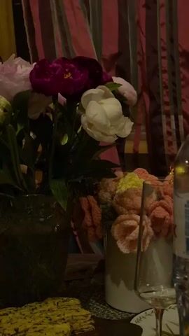 The video is a close-up shot of a table with floral arrangements and food. In the foreground, a plate of what appears to be crackers or flatbread topped with a yellow spread is visible. To the right, a wine glass and a partially visible bottle of water are placed. In the background, two vases of flowers are prominent. The vase on the left contains a large bouquet of peonies in shades of pink, purple, and white. The vase on the right holds a smaller arrangement of crocheted flowers in various pastel colors, including peach, pink, and yellow. The backdrop features striped curtains in shades of pink and brown. The lighting is dim, creating a moody atmosphere.