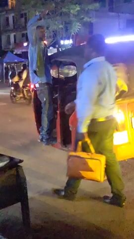 The video shows a man standing on the side step of a red and black auto rickshaw at night. He's wearing a light blue hooded sweatshirt, dark jeans, and dark shoes. He appears to be looking up, with one arm raised. Another man in a light blue button-down shirt and dark pants is walking away from the rickshaw, carrying a brown leather briefcase. The street is illuminated by streetlights and the rickshaw's headlights. Other vehicles, including motorcycles and other rickshaws, are visible in the background. There is also a building and some trees in the background.