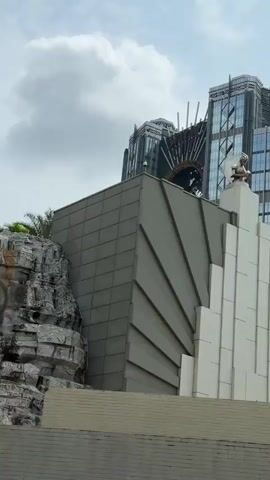 The video shows a modern architectural scene with a focus on a building adorned with a statue and an Art Deco design. The foreground features a gray and white building with a striking white spire. A statue sits on top of the spire. The gray walls of the building are decorated with a fan-like pattern. In the background, there is a tall, modern skyscraper with glass windows. A golden circular structure is visible behind the skyscraper, perhaps part of another building or architectural feature. A water slide is also visible on the building. The sky is overcast. The camera pans slightly, providing different views of the buildings and structures.