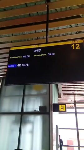 Here's a description of the video:

The video shows an airport boarding gate area. A flight information display shows the destination as Jaipur, with both scheduled and estimated departure times listed as 09:50. The flight number is Indigo 6E 6978.  The gate number is 12. 

The camera then pans to show a large window looking out onto a tarmac or landing area. Two men are visible near the window, though their activities are not clear. The ceiling has a modern slatted design. A yellow sign with an upward arrow is visible near the exit.