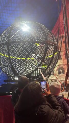 The video shows a family of three (a child in a gray hat, a dark-haired woman in a black jacket, and an unidentifiable man) sitting in front of a stage featuring a large, spherical cage-like structure. A motorcyclist inside the cage is performing stunts. The woman holds a smartphone, recording the event as the motorcyclist maneuvers around the inside of the sphere. The surrounding environment includes a blue, tent-like ceiling, red curtains, and background scenery depicting a classical building.