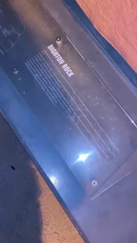 The video shows a close-up view of a plaque with text that reads "DIGHTON ROCK." The plaque is mounted on a dark gray surface, possibly a bench or a display case. The background is a rough, orange-colored wall or structure. The lighting suggests it might be nighttime or indoors with artificial light. The plaque appears to be made of clear acrylic with the text etched or printed on it. Some dust or smudges are visible on the surface of the plaque.