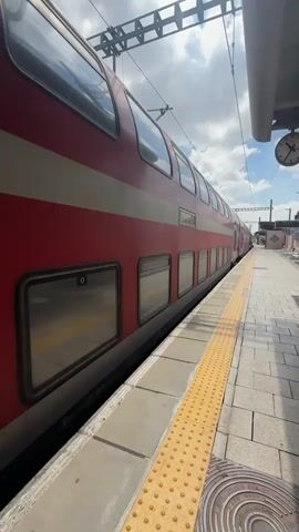 The video captures a view of a double-decker train at a train station platform. The train is primarily red with a white stripe running horizontally along its length. The platform has a yellow tactile paving strip along its edge, indicating a safety feature for visually impaired individuals. The train is stationary, suggesting it is either waiting for passengers or has just arrived.

The setting appears to be an outdoor train station on a bright day. The sky is visible, filled with scattered clouds, and the sunlight casts shadows, indicating it's likely daytime. Overhead, electrical wires for the train are prominent. In the distance, a clock is visible on a pole, but its face is too small to read the exact time.

There are no people visible in the frame. The focus is on the train and the platform. The overall mood is calm and observational. A small piece of text is visible on the side of the train near the bottom, appearing to be numbers, possibly a train car identification number. Further text is visible on the side of the train with the number "2267". The location context provided is Hod HaSharon, Israel.