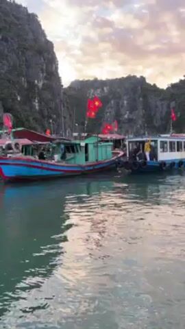 The video shows a scenic view of a harbor or bay with boats docked against a backdrop of steep, rocky islands under a colorful sky. The water is relatively calm, reflecting the colors of the sky. In the foreground, there are two main boats: one painted in teal and red with Vietnamese flags and the other is a white and blue tour boat. People are seen boarding the white and blue boat. The Vietnamese flags are on both boats. The sky features a mix of light blue and soft pink and orange hues, indicating either sunrise or sunset. The overall setting is calm and picturesque.