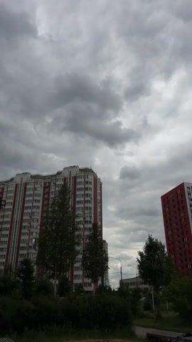 The video shows a view of the sky above two residential buildings. The sky is filled with dark, heavy clouds, indicating an overcast or stormy day. The buildings are tall, multi-story structures with visible windows and balconies. Trees and some foliage are visible in the foreground, obscuring the lower portions of the buildings and the ground. The camera appears to be stationary, capturing the clouds moving slowly across the sky.