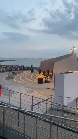 The video depicts an outdoor evening concert held in Vladivostok, Russia, likely at a waterfront venue. The scene is illuminated by numerous candles arranged in concentric circles on the ground, creating a warm and inviting atmosphere. In the center of these circles, a small musical ensemble is performing on a stage. Several people are seated on the ground facing the performers, while others stand behind them. The audience is gathered along the coastline, suggesting the concert is taking place near the sea.

In the background, the ocean stretches out towards the horizon, with ships visible in the distance. The sky is overcast with dramatic clouds, indicative of either dusk or a stormy weather approaching. The architecture of the venue is modern and angular, with concrete structures and metal railings framing the performance area. A large, white rectangular structure is also present, possibly a screen or a decorative element. The overall mood is serene and reflective, amplified by the soft glow of the candles and the tranquil setting. There is no visible text in the video.