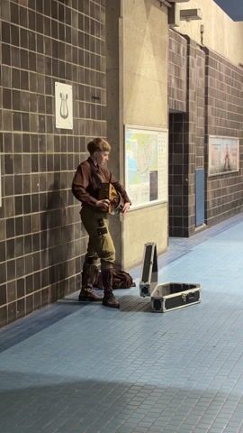 Here is a description of the video:

A full-shot captures a young adult standing in a tiled corridor, adjusting the straps of an instrument. The person is dressed in a brown button-down shirt, khaki-colored pants, and brown boots, appearing to be in their early twenties.  Two black cases, likely for instruments, are placed on the blue tiled floor near the person. The corridor features black tiled walls on one side and a combination of black tiles and concrete pillars on the other.  A map is visible on the wall behind the person.  The setting appears to be an underground public space like a subway or train station.  The person is holding a tan stringed instrument and adjusting its position.