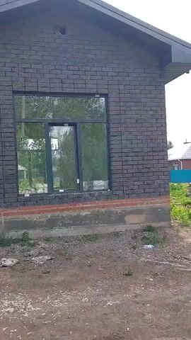 The video shows a tour of a brick house under construction. The house has large windows and a dark gray door. There's a blue barrel and some construction materials near the front entrance. The yard is still under construction with dirt and some sparse grass. In the background, there's another similar house and glimpses of the neighborhood. The camera pans from the front of the house across to the door.