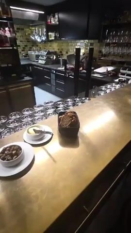 A bar counter is shown with various items placed on it. In the foreground, there are two small white plates. The plate on the left contains a bowl filled with what appears to be olives or small pickled items. The plate to its right holds a bowl of butter with a knife resting on it. In the center of the counter, a dark basket contains slices of bread. Behind the counter, rows of clean glassware are neatly arranged. The background reveals a well-equipped kitchen area with stainless steel appliances, shelving stocked with bottles, and tiled walls. The overall scene suggests a bar or restaurant setting, possibly preparing for service or having just finished. There are no people visible in the video.