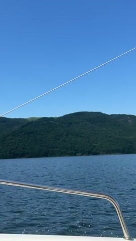 The video captures a view from the deck of a boat moving across a body of water. A clear blue sky dominates the upper portion of the frame. In the distance, a large, tree-covered hill or mountain rises from the water's edge. The water itself is a deep blue and appears slightly choppy, indicating movement. In the foreground, a portion of the boat's railing, made of polished metal, is visible along the bottom edge of the frame. A white rope or cable is stretched diagonally across the sky. The overall impression is one of a scenic boat ride on a clear day.