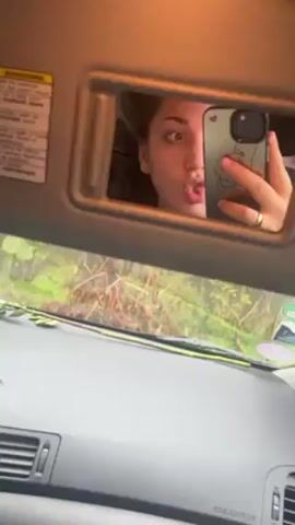 A young woman is sitting in a car and taking a selfie using the vanity mirror on the sun visor. The camera is positioned in front of the dashboard, capturing the interior of the car as well as the woman's reflection. Outside the windshield, we see a green, wooded area. The woman has her mouth puckered in a kissy face expression. She is holding an iPhone with a decorative case. The dashboard is gray and has air vents. A warning label is visible on the sun visor.