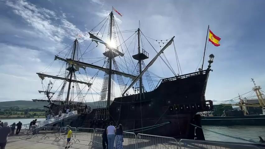 The video shows a large, dark-colored, historical-style sailing ship docked at a pier. A Spanish flag flies from the stern of the ship. The ship has multiple masts and rigging, suggesting it is a replica of a galleon or similar vessel. There are people on the pier, some standing and observing the ship, while others appear to be boarding or disembarking via a ramp. The sky is blue with some scattered clouds, and the sun is visible, shining brightly behind the ship's masts, creating a lens flare effect. The water is calm and reflects the sky and ship. Another, more modern-looking ship is partially visible in the background to the right. The environment appears to be a harbor or marina on a sunny day.