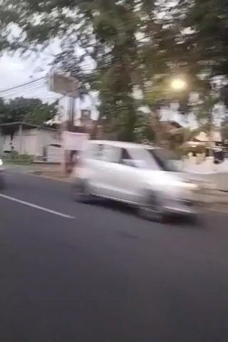 The video shows a side-view of a road with vehicles driving on it. In the foreground, a motorcycle's mirror is visible. The road is paved with white lane markings. On the right side of the road, there are trees, houses, and utility poles. On the left side of the road, there are more trees and buildings. Several vehicles are seen including a black SUV, a white SUV, a bus, and a yellow truck. The sky is overcast. The video appears to be shot from another moving vehicle, possibly the motorcycle.