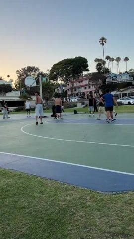 The video shows a basketball court with several people playing. The court is green and blue with white lines. There are trees and buildings in the background, and the sky is clear and blue. Some of the people are wearing athletic clothing. The camera is positioned low, looking up at the players. The video appears to be taken during the day.