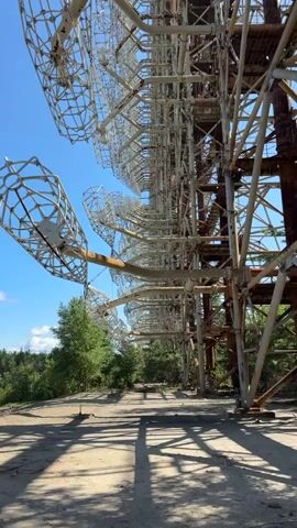 This video captures a low-angle view of a large, abandoned radar installation against a bright blue sky. The structure, made of metal beams and lattice-like parabolic dishes, extends vertically and horizontally. The metal is rusted in places, indicating age and disuse. In the foreground, a dirt path with shadows cast by the radar structure stretches into the distance. Green trees and foliage border the path and the base of the radar array, suggesting an outdoor, possibly overgrown, environment. The camera pans slightly, offering a detailed look at the complex, repetitive design of the radar components. There are no people or animals visible in the footage.