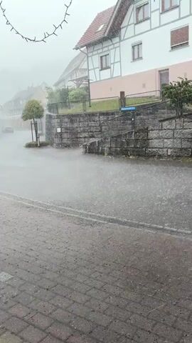 The video captures a village street in Forbach, Germany, during a heavy downpour. The scene is dominated by the weather, with rain falling intensely, creating a misty and wet atmosphere. Water is visible splashing on the paved road and sidewalk, and there appears to be significant spray from vehicles.

The setting is a quaint European village, characterized by traditional half-timbered houses. One prominent house in the foreground has a white facade with timber framing and a red-tiled roof. Another house on the left is painted red and also features timber accents. Several cars are parked along the street, and a small tree stands by the roadside, looking somewhat battered by the rain. A stone wall with a metal fence runs along the side of the street, and a small staircase leads up from the road. Overhead, decorative string lights are visible, seemingly unaffected by the rain.

The time of day appears to be daytime, though the heavy cloud cover and rain obscure direct sunlight, creating a muted light. The overall mood is one of a sudden, intense rain shower, possibly a summer storm, creating a dynamic and somewhat dramatic natural event in the otherwise peaceful village setting.

Visible text includes a street sign with "Mittelweg 12 a" on it. There's also a house number "6" visible on the white house in the foreground.