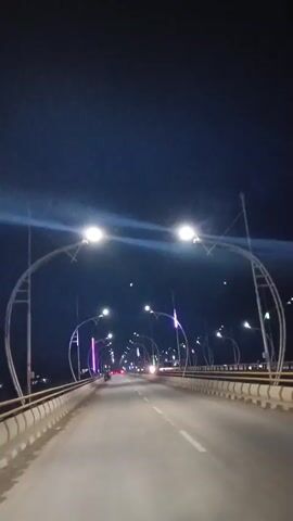 A first-person perspective video shows a view from a moving vehicle at night on a bridge. The bridge is illuminated by streetlights and decorative lights. The road is paved and has white lane markings. On either side of the road are railings and sidewalks. In the distance, other vehicles can be seen, including motorcycles. The sky is dark blue, suggesting it is either dusk or night. The overall impression is that of a commute or drive across a well-lit bridge.